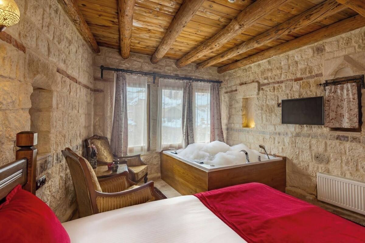 A rustic room with a stone wall, a wooden ceiling, a large bed with a red blanket, a wooden bathtub filled with bubbles, a wooden chair, and a small table with a lamp.