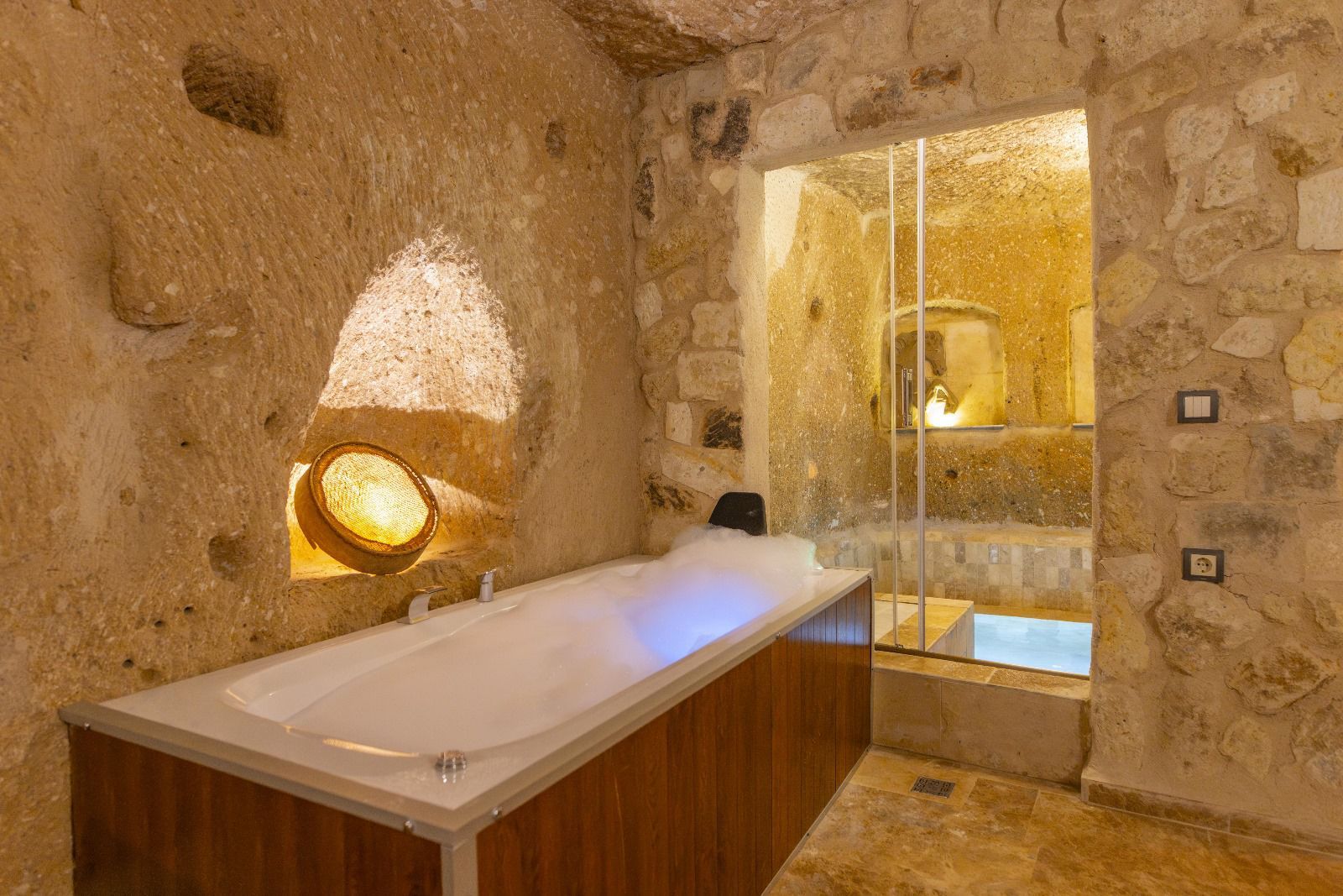 Casa de banho com paredes de pedra, toucador de madeira, cabine de duche em vidro e luminária fixada na parede.