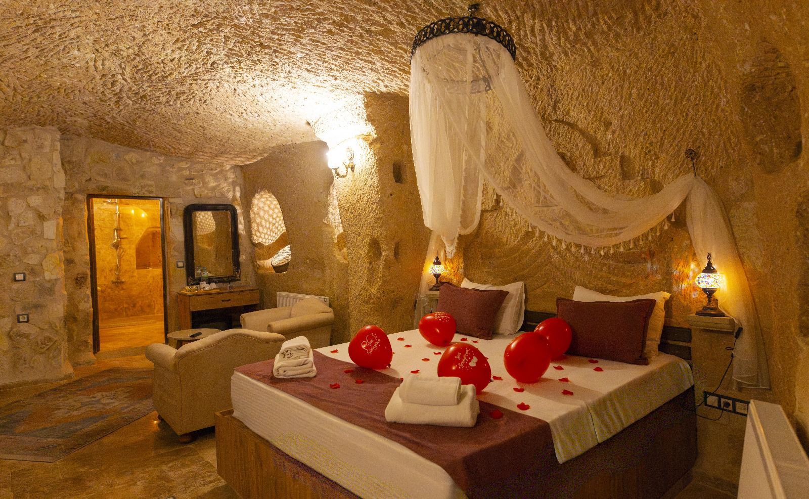 A cozy bedroom with a stone wall, a bed with red balloons, a vanity with a mirror, and a shower area.