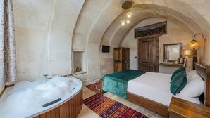 A room with a wooden bed, a bathtub, and a rustic stone wall. - Zula Cave House (Ürgüp)