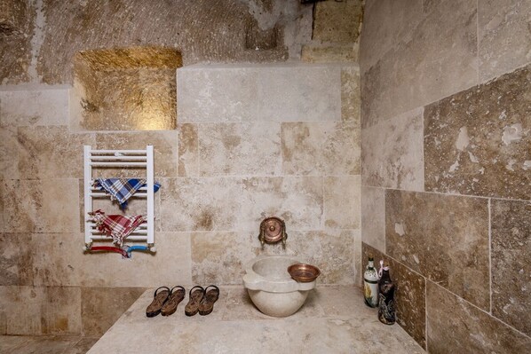 A bathroom with a bathtub, a towel rack with a plaid towel, a pair of sandals, and a copper basin.
