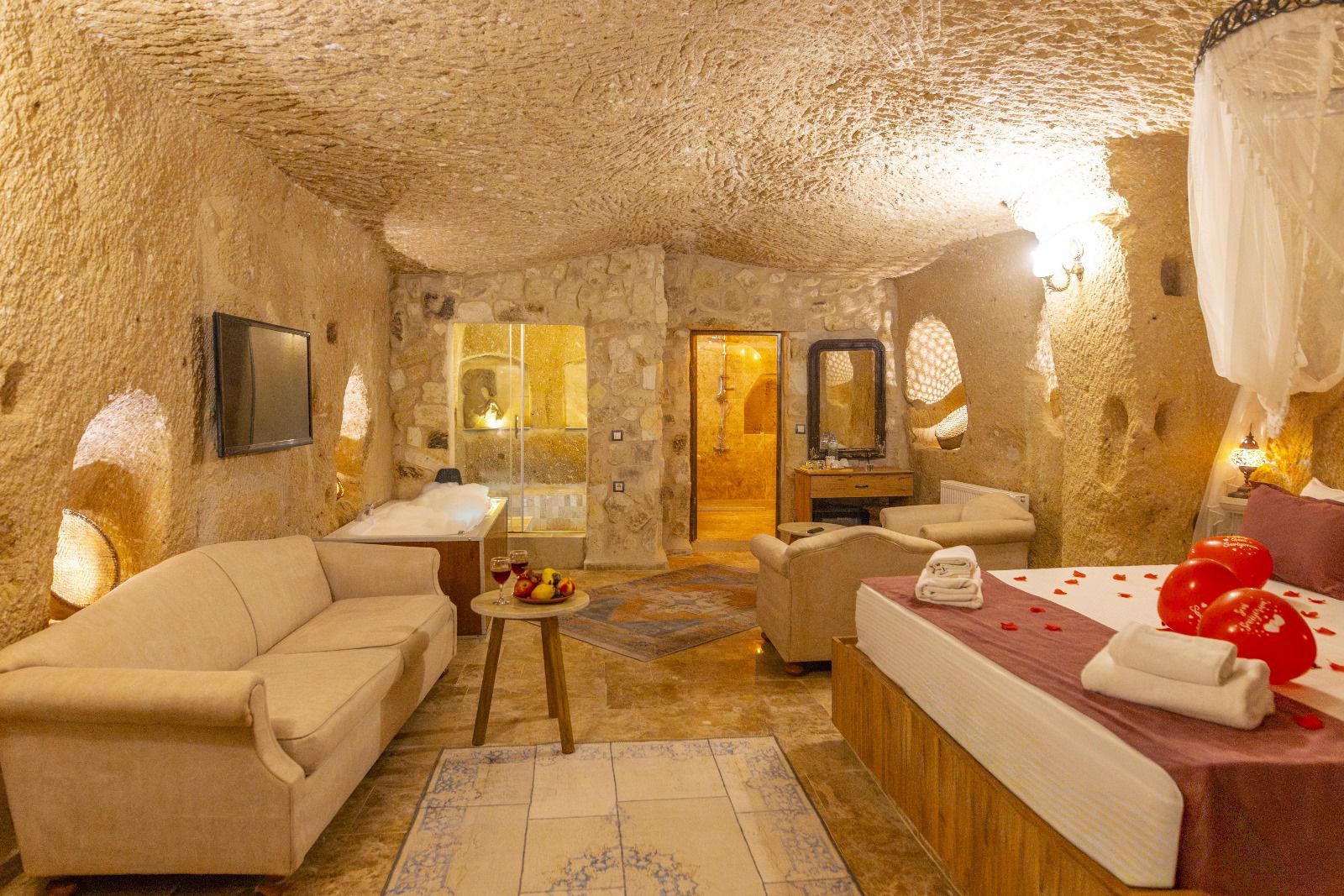 Quarto com parede de pedra, banheira, sofá, mesa com frutas e cama com balões vermelhos.