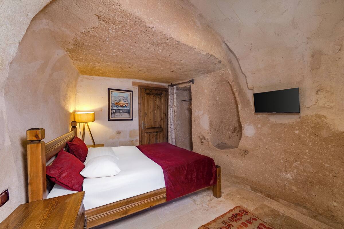 A bedroom with a wooden bed, a red blanket, a wooden headboard, a nightstand, a lamp, a framed picture on the wall, and a television mounted on the wall.