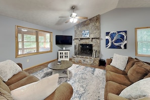 A living room with a stone fireplace, a ceiling fan, a flat-screen TV, and a large window with blinds. - Carefree Cove by Taylor Made Deep Creek (Swanton)