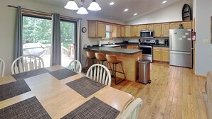 A modern kitchen with wooden cabinets, a central island, and a dining area with a table and chairs. - Carefree Cove by Taylor Made Deep Creek (Swanton)