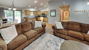 A modern living room with brown sofas, a patterned rug, and a kitchen in the background. - Carefree Cove by Taylor Made Deep Creek (Swanton)