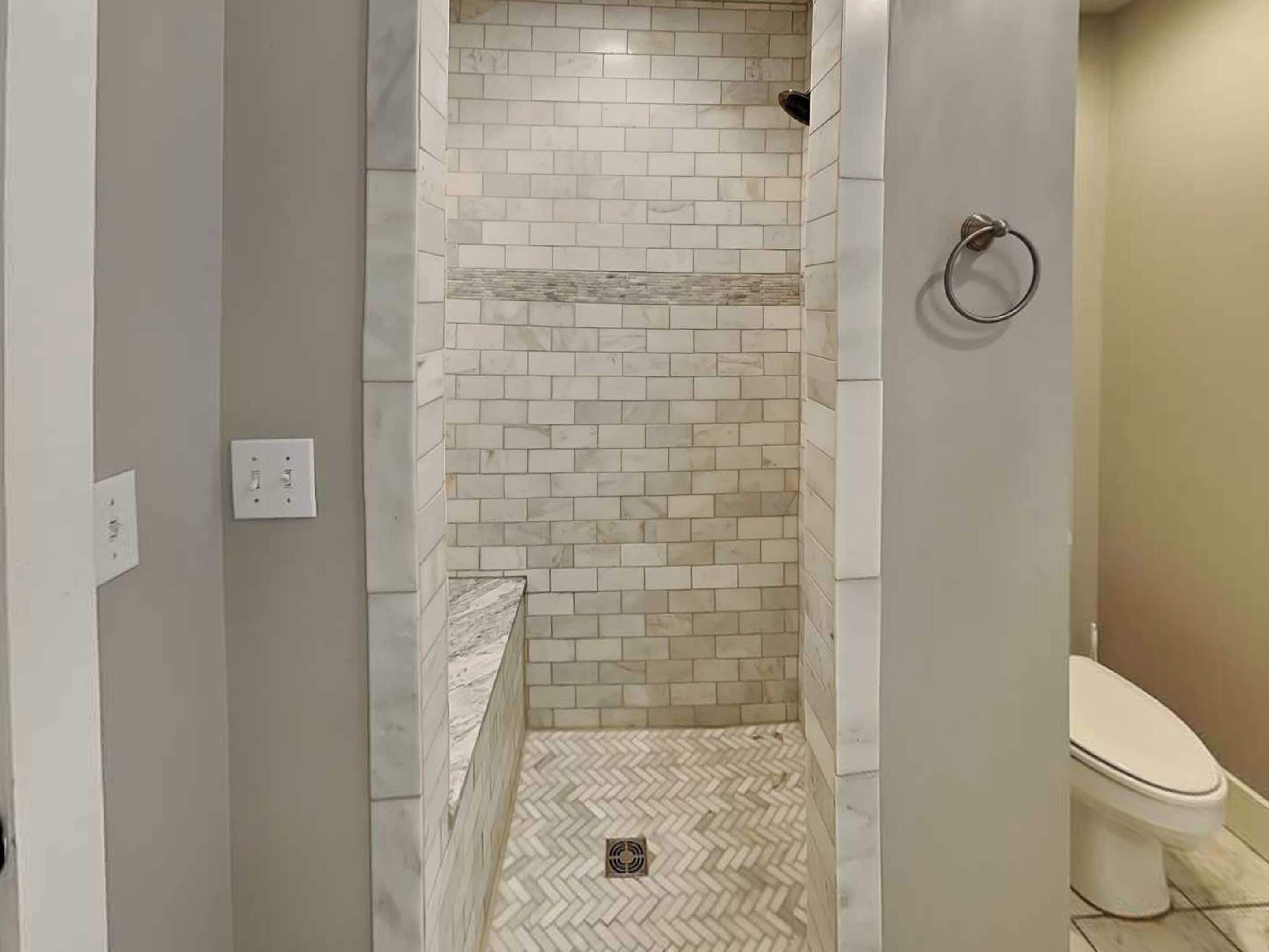 Un baño con zona de ducha revestida de baldosas blancas y grises, suelo con diseño de espiga y un inodoro.