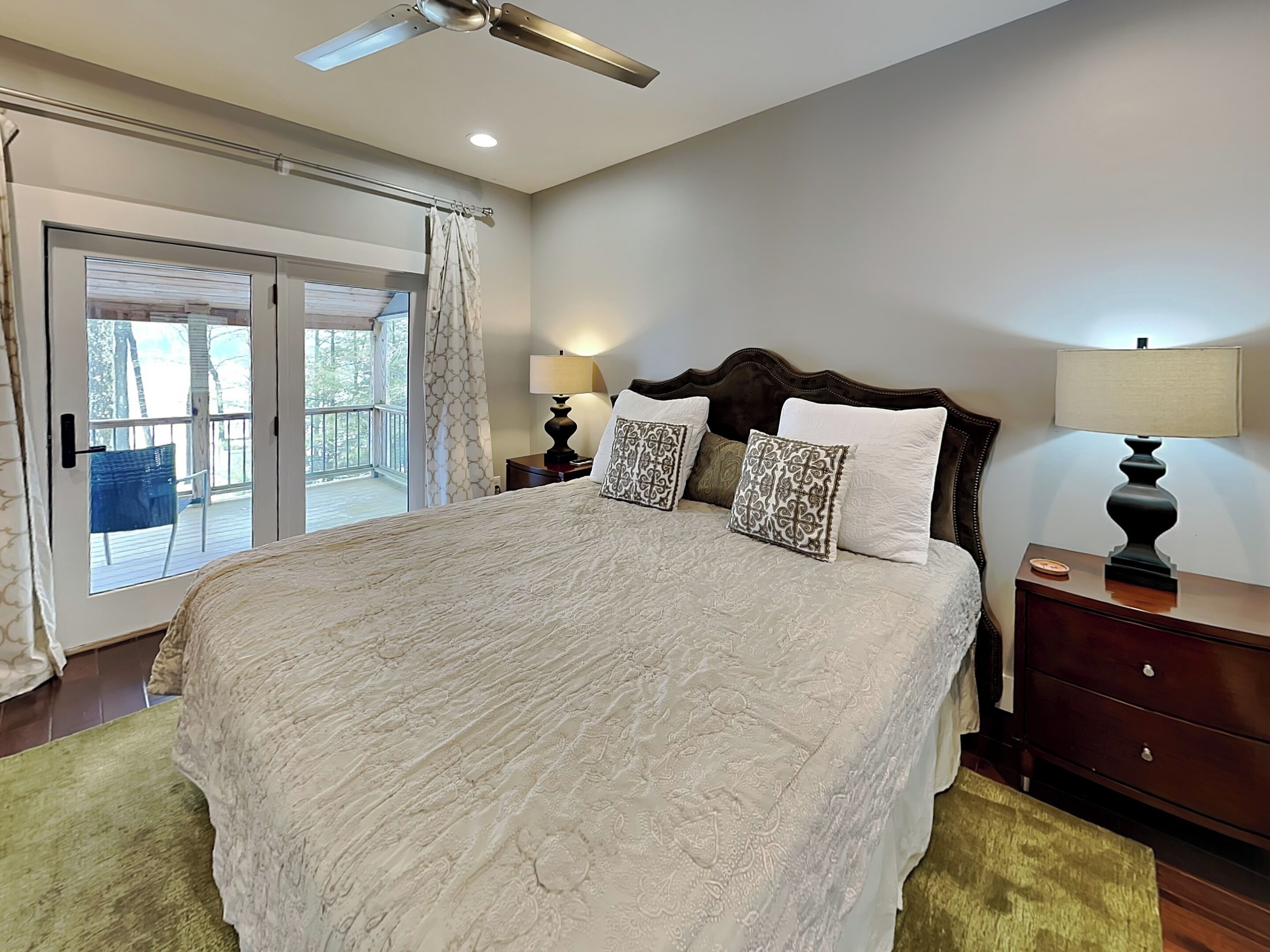 A spacious bedroom with a large bed, a wooden headboard, two bedside tables with lamps, and a sliding glass door leading to a balcony.