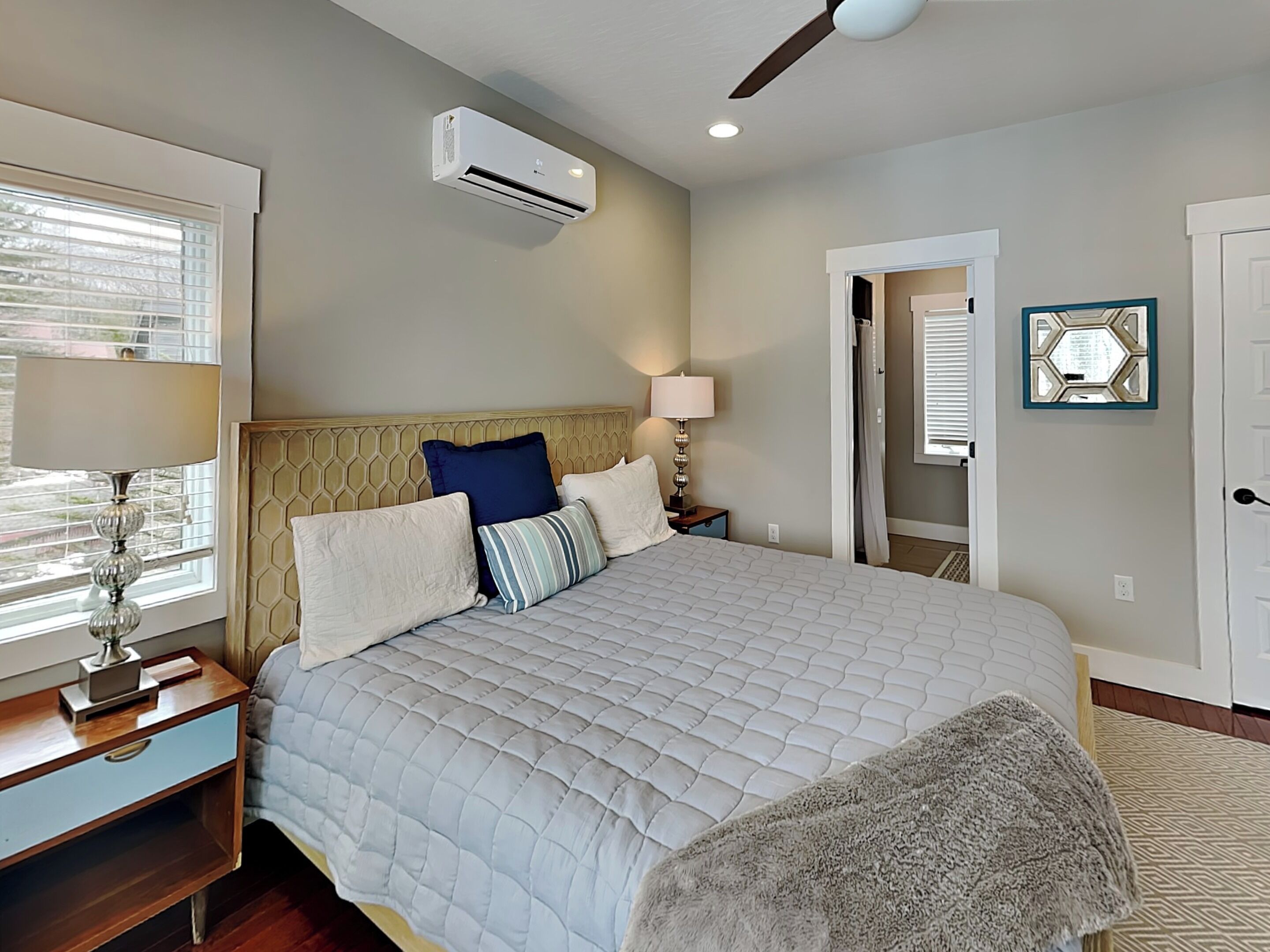 A bedroom with a bed, bedside lamps, an air conditioning unit, a ceiling fan, and a door leading to another room.