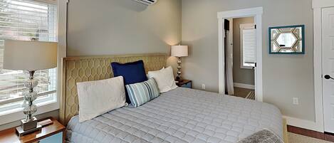 A bedroom with a bed, bedside lamps, an air conditioning unit, a ceiling fan, and a door leading to another room.