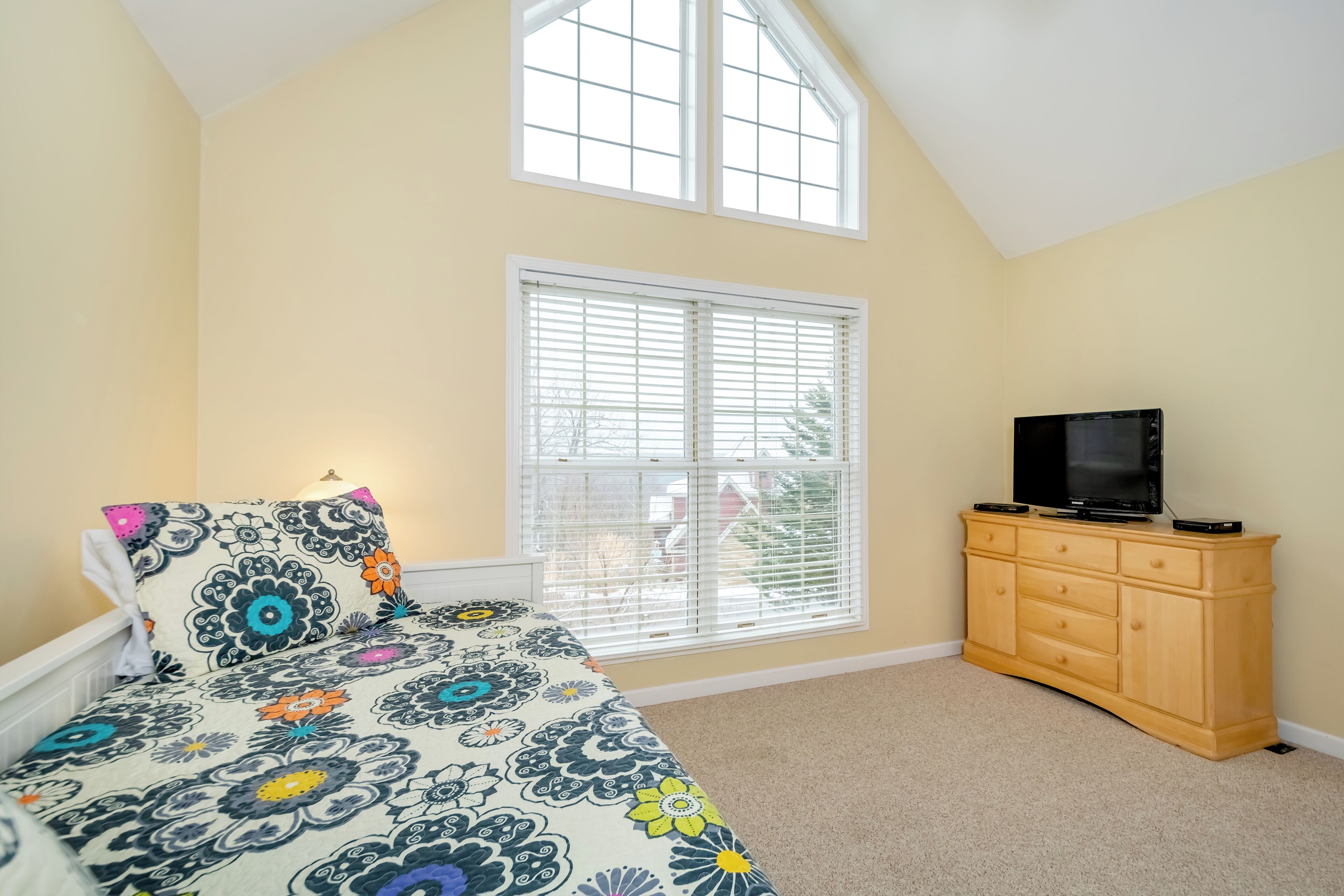 A bedroom with a bed, a TV on a wooden cabinet, and a window with blinds.