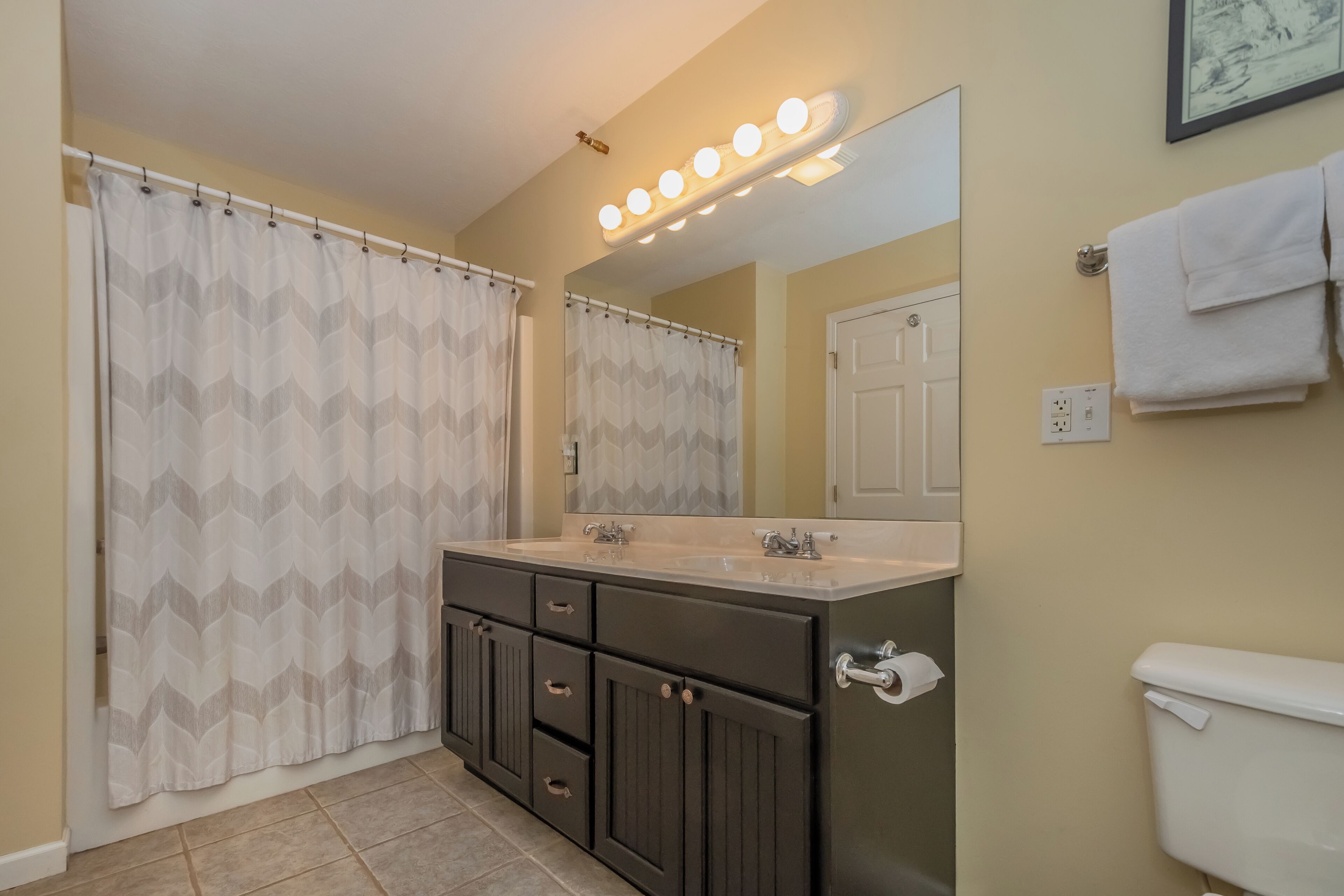 A bathroom with a vanity, double sink, mirror with lights, shower curtain, and a toilet.