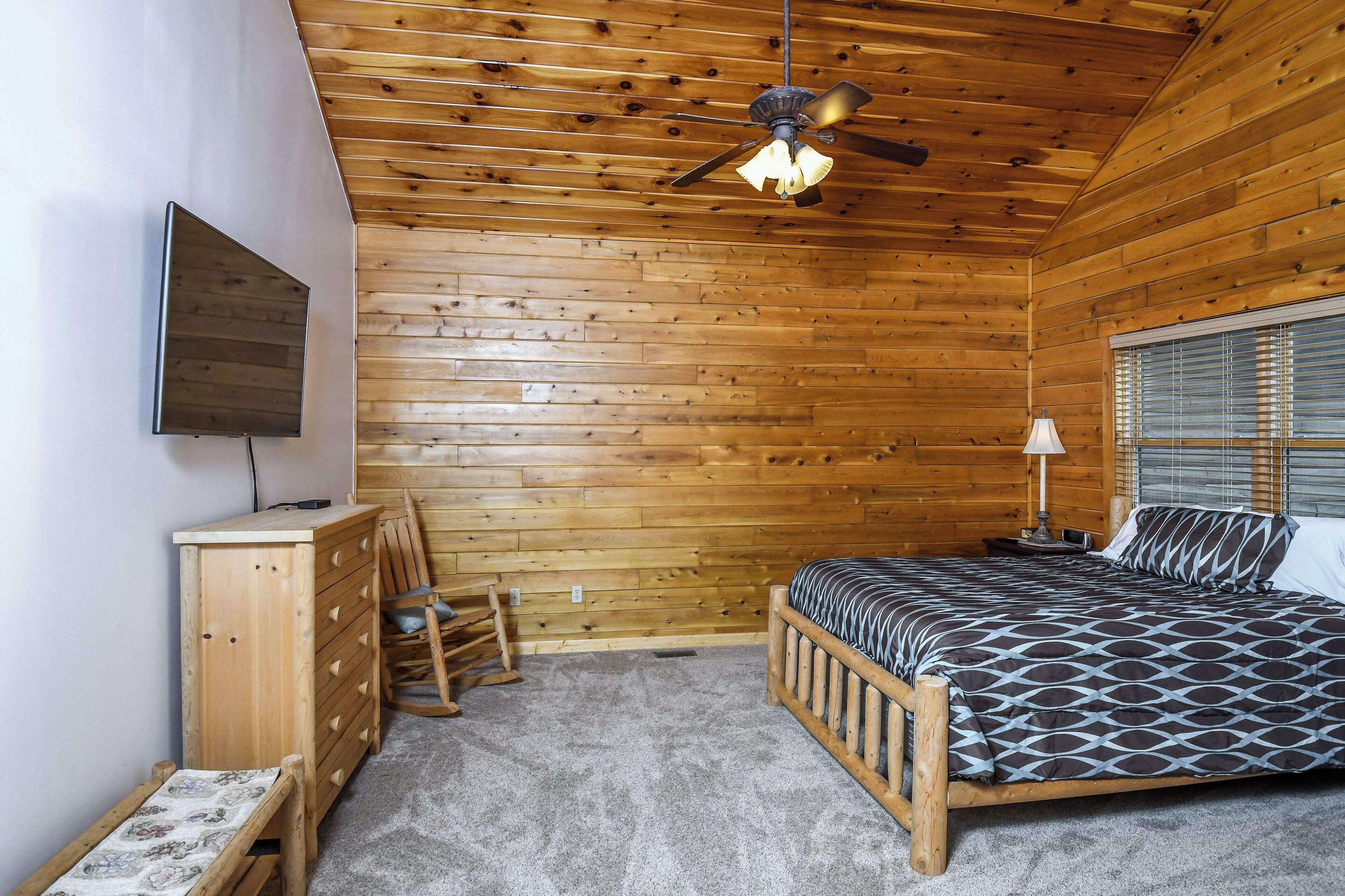 Un dormitorio con techo de madera, una cama con ropa de cama estampada, una mesita de noche, una lámpara, un aparador y un televisor montado en la pared.