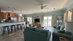 A spacious living room with a green sofa, a coffee table, and a TV. There is a kitchen area with wooden cabinets and a bar with stools.