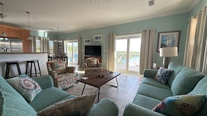A spacious living area with a sofa set, a coffee table, and a television. There is a balcony with a view, and the room has a kitchen area with cabinets and a refrigerator.