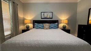 A neatly made bed with a patterned bedspread, two bedside lamps, a framed painting above the bed, and a window with blinds on the left.
