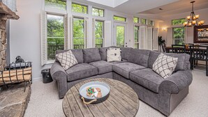 Un salon spacieux avec un canapé d’angle gris, une table basse en bois et de grandes fenêtres donnant sur un paysage verdoyant.