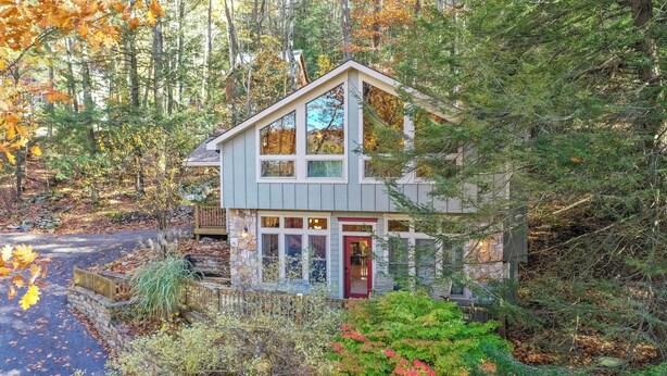 Une cabane moderne à deux étages, dotée de fondations en pierre, de grandes fenêtres et d’une porte rouge, entourée d’une forêt dense et d’un feuillage automnal.