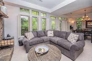 A spacious living room with a gray sectional sofa, a wooden coffee table, and large windows offering views of greenery.