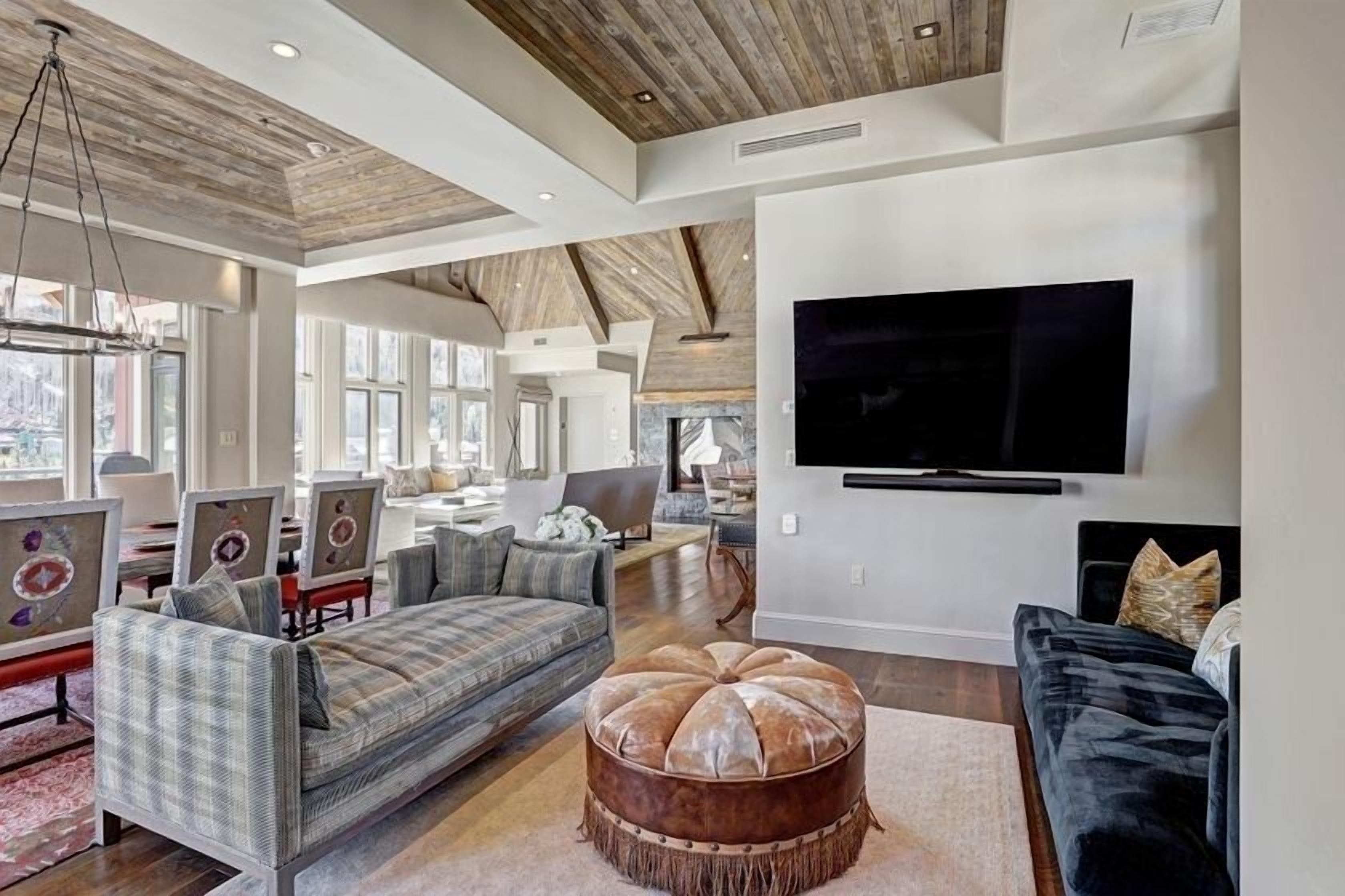 A spacious living room with a large flat-screen TV mounted on the wall, a wooden ceiling, and a cozy seating area with a round ottoman.