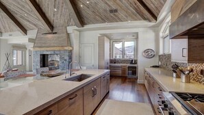 A spacious kitchen with a stone fireplace, wooden cabinets, and a large island with a sink. - Luxury Mountain-Side Penthouse with Stunning Views! by RedAwning (Vail)
