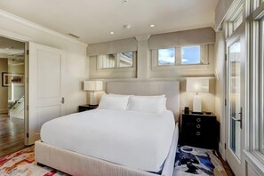 A large bed with white linens, two bedside tables with lamps, a patterned rug, and a view of a hallway with a staircase. - Luxury Mountain-Side Penthouse with Stunning Views! by RedAwning (Vail)