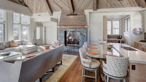 A spacious living area with a stone fireplace, a modern sofa, a dining table with chairs, and a kitchen area with a large window. - Luxury Mountain-Side Penthouse with Stunning Views! by RedAwning (Vail)
