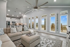 Un salon spacieux, doté d’un grand canapé d’angle, d’un ventilateur de plafond et offrant une vue sur l’extérieur grâce à de grandes fenêtres.