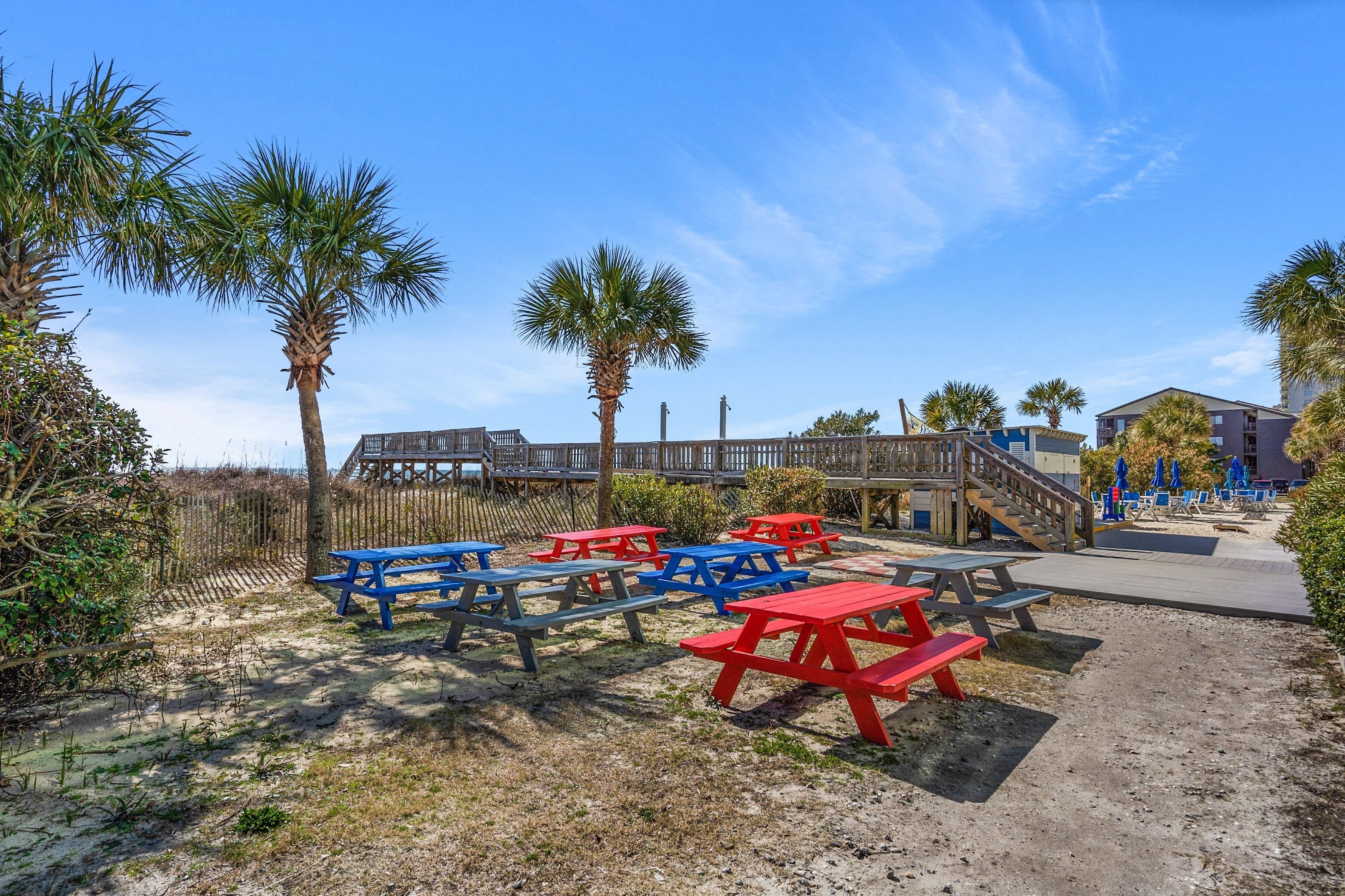 A beachside area with picnic tables, palm trees, and a wooden boardwalk leading to a building.