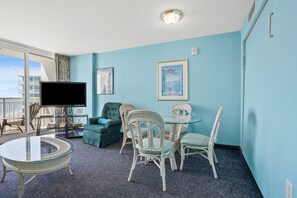 A room with a large window, a flat-screen TV, a round glass table, and a seating area with wicker chairs and a sofa. - Avista Resort 1503 1 Bedroom Condo by RedAwning (North Myrtle Beach)
