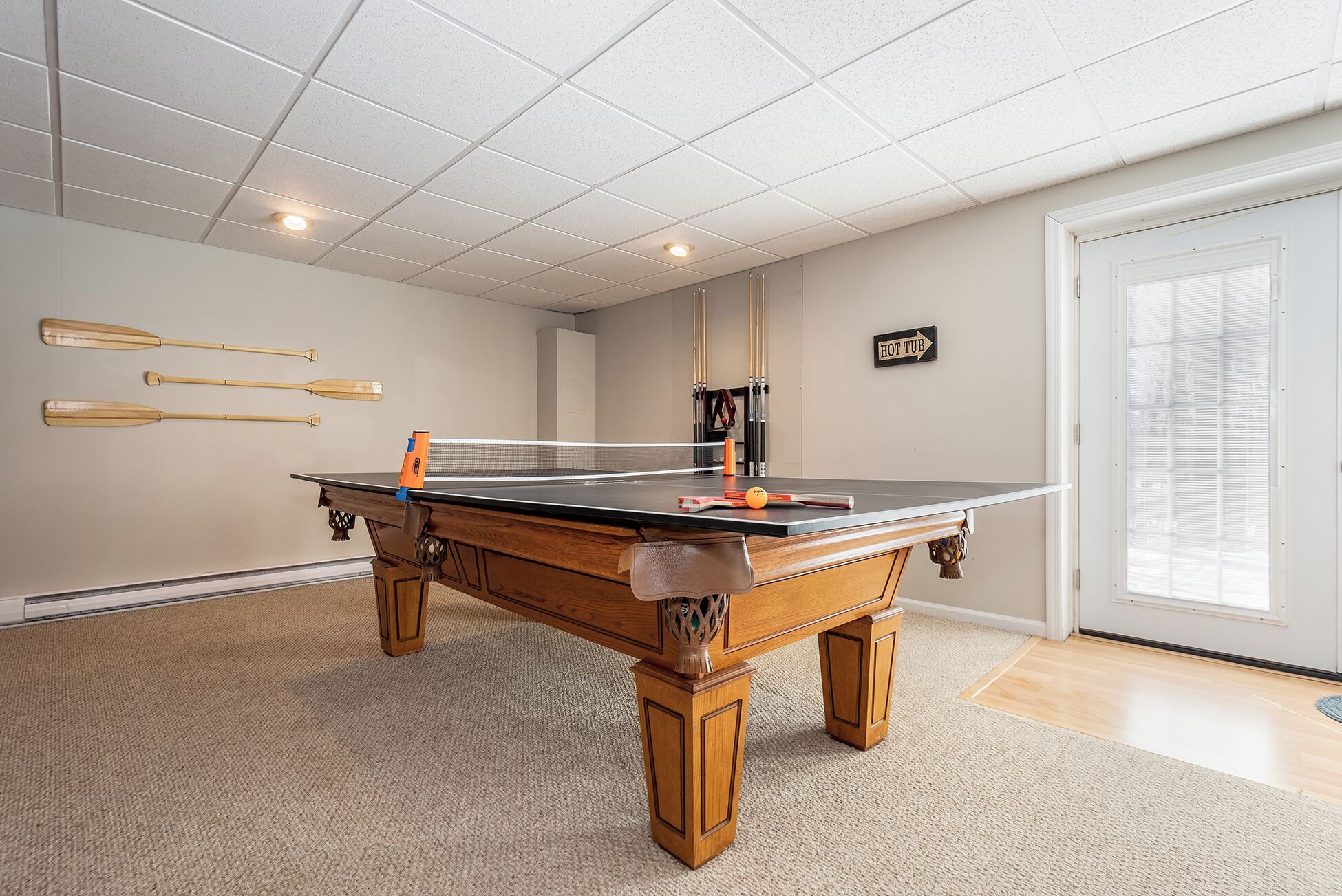 Une table de ping-pong dans une pièce dont le sol est recouvert d’une moquette, avec une porte et une fenêtre équipée de stores.