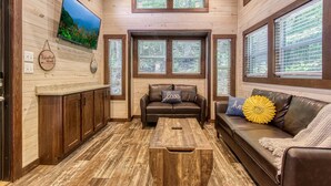 Una sala de estar acogedora con detalles de madera, un televisor de pantalla plana en la pared, un sofá de cuero marrón con cojines decorativos y una mesa de centro.
