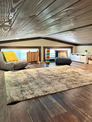 An attic room with wooden flooring, a large rug, two armchairs, a wooden play structure, and a window with blinds. - Smoky Mtn Tiny Home+Hot Tub, Fire Pit & Views+2 miles to the Smokies (Sevierville)