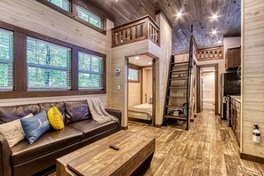 A cabin interior with a loft bedroom, a living area with a sofa and coffee table, and a kitchen area.