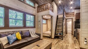 Interior de una cabaña con habitación en altillo, un sofá con cojines, una mesa de centro de madera y una zona de cocina.