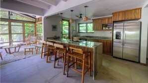 A spacious kitchen with a large island, wooden bar stools, a stainless steel refrigerator, and large windows offering a view of greenery.