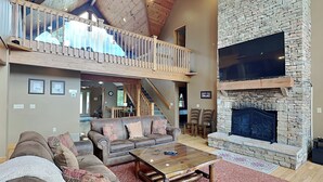 A spacious living area with a high ceiling, a stone fireplace, and a balcony overlooking the interior. - Celebration at Deep Creek by Taylor Made Deep Creek (Oakland)