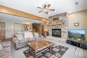 A cozy living room with a stone fireplace, a wooden ceiling fan, and a television. - Celebration at Deep Creek by Taylor Made Deep Creek (Oakland)