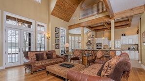 A spacious living room with a high ceiling, wooden beams, and a large dining area with a kitchen in the background. - Champagne Taste by Taylor Made Deep Creek (Oakland)