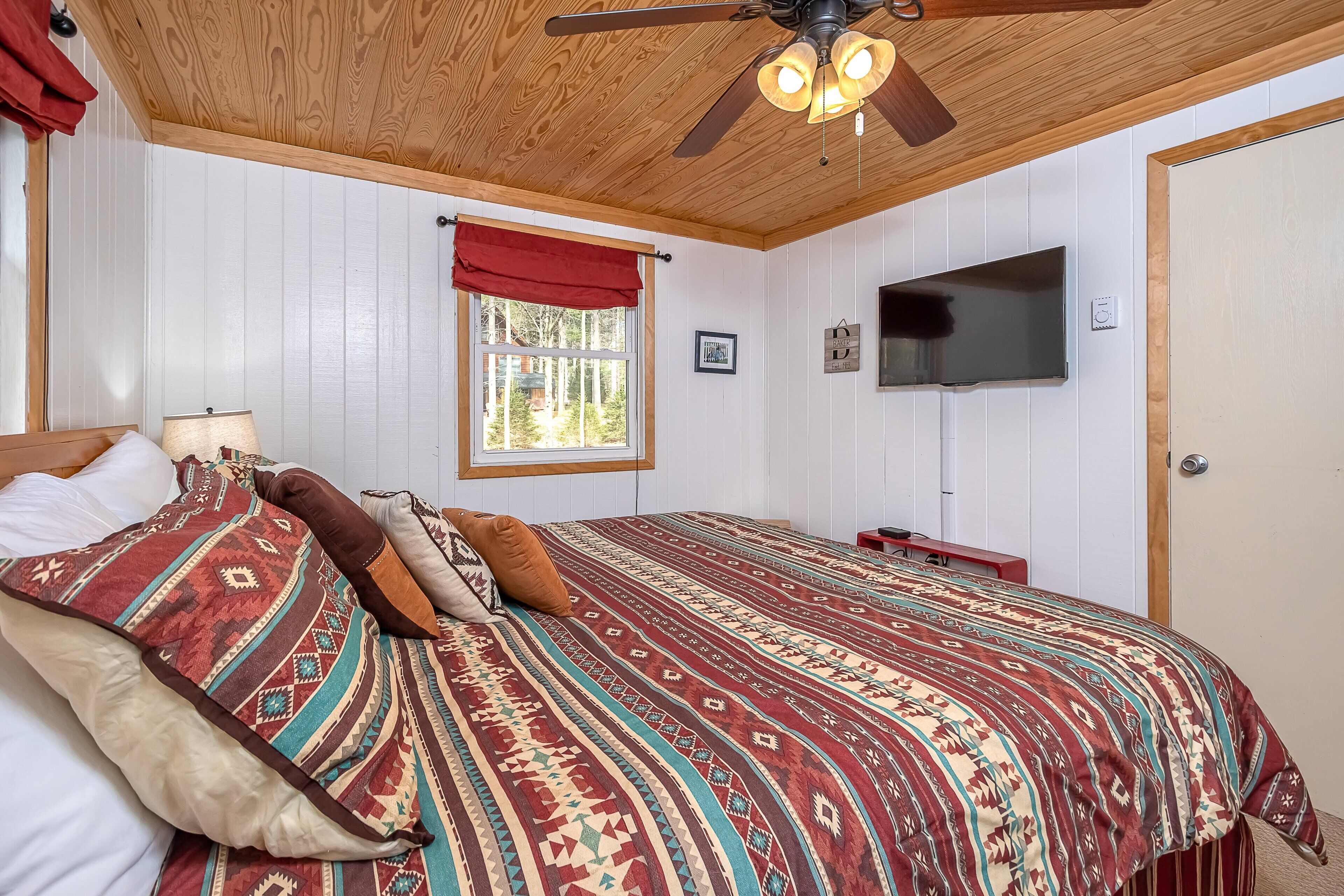 Ein Schlafzimmer mit Holzboden, einem Bett mit gemusterten Betttextilien, einem Fenster mit roten Vorhängen, einem Wand-Fernseher und einem Deckenventilator.