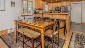 Un interior de madera con una mesa de comedor de cristal, sillas, una cocina con armarios de madera y una alfombra estampada.