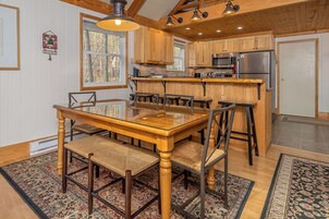 A wooden interior with a glass dining table, chairs, a kitchen with wooden cabinets, and a patterned rug.