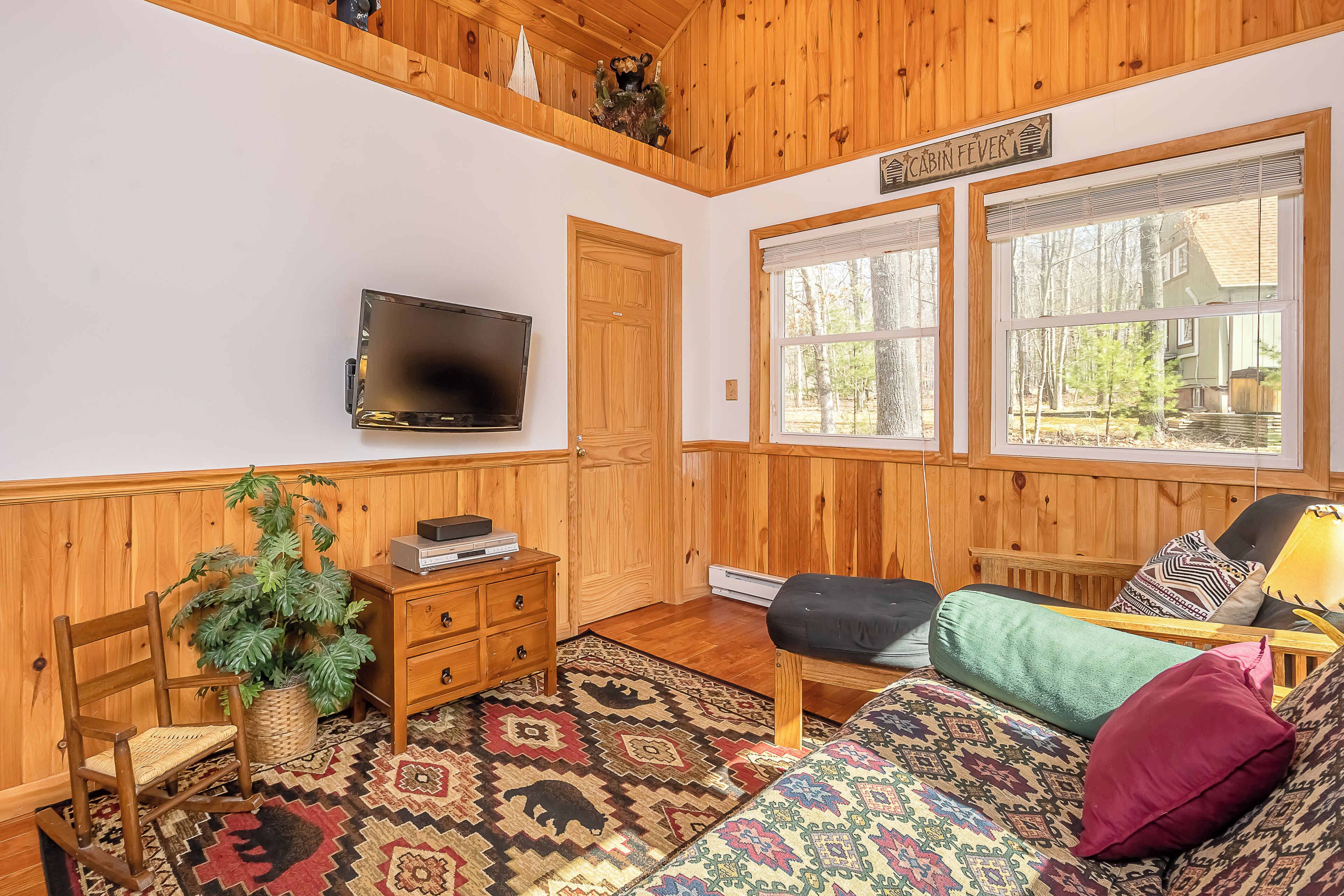 A cabin-style room with wooden walls and ceiling, a television mounted on the wall, a bed with patterned bedding, a chair, a small table with a plant, and a window with a view of the outside.