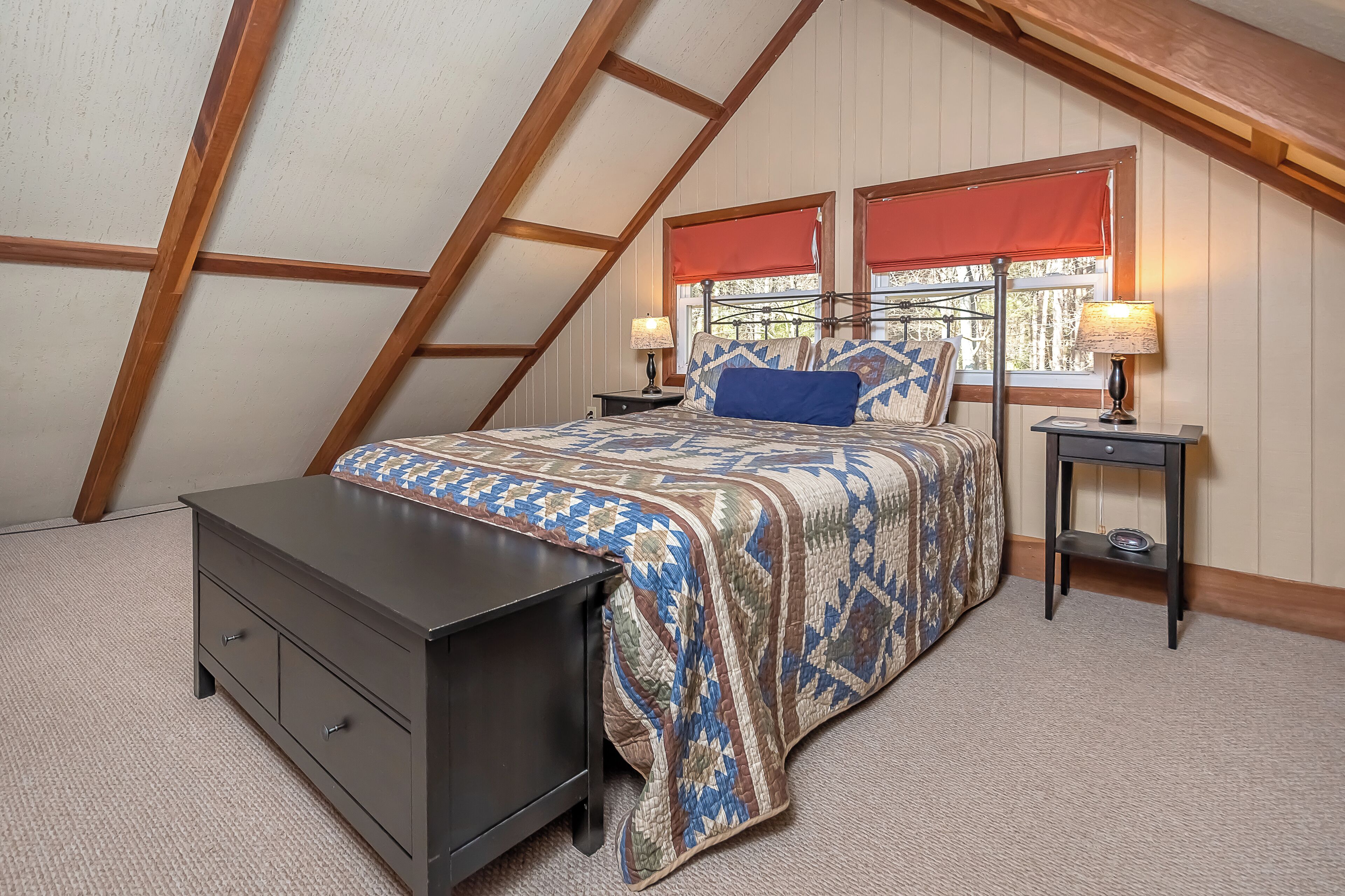A bedroom with a sloped ceiling, a bed with a patterned blanket, two bedside tables with lamps, and a dresser.