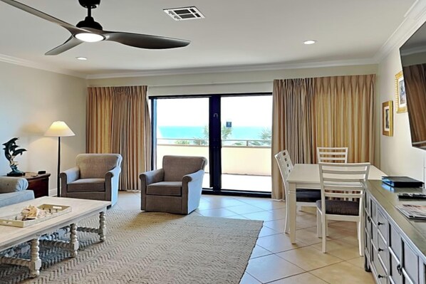 Ein Wohnzimmer mit Deckenventilator, einer Couch, einem Couchtisch und Blick auf den Strand durch große Fenster.