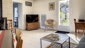 A living room with a TV, wooden furniture, a glass coffee table, and a view through a glass door. - VILLA LA COTINIERE (Saint-Pierre-d'Oléron)