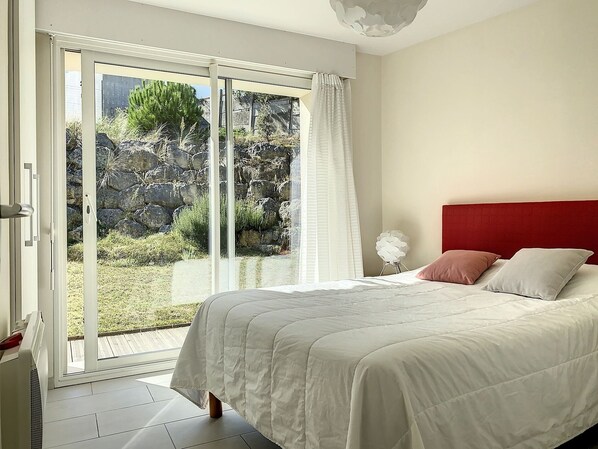 Quarto com cama, cabeceira vermelha e vista para uma área externa rochosa através de uma porta de vidro deslizante.