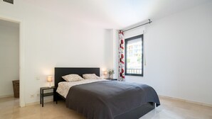 Un dormitorio con cama, mesitas de noche, una lámpara y una ventana con vistas a edificios.