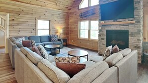 A log cabin interior with a stone fireplace, a large sofa, a coffee table, and a television mounted on the wall. - Cj's Mountain Lodge by Taylor Made Deep Creek (McHenry)