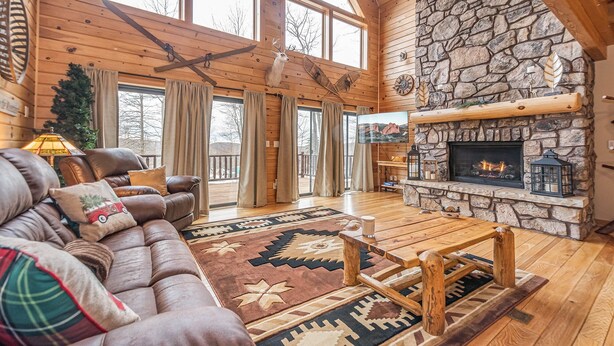 A cozy log cabin interior with a stone fireplace, leather sofa, and a large window overlooking a snowy landscape. - Colorado Dreamin' by Taylor Made Deep Creek (McHenry)